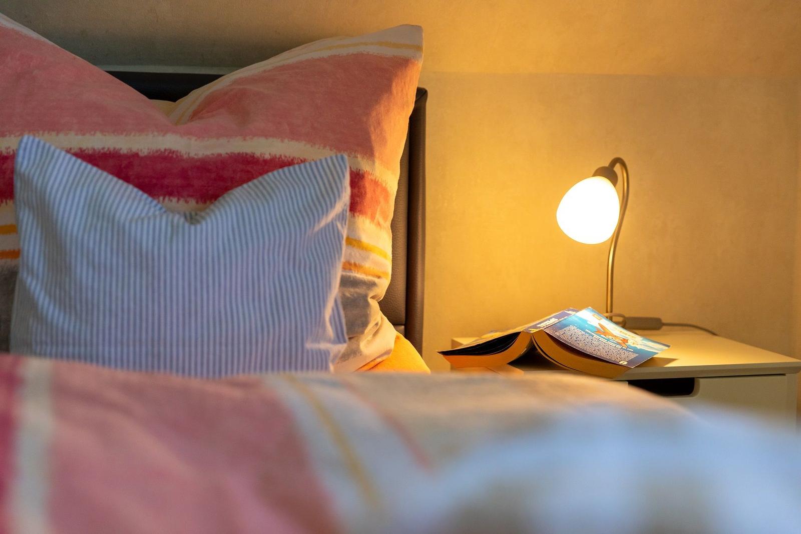 Bettkopf mit Kissen und Nachttisch mit Lampe und Buch.