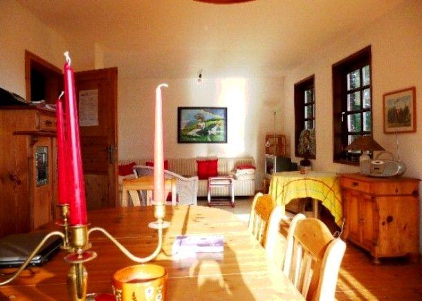 Esszimmer mit Holztisch, Stühlen und Kerzenhalter. Im Hintergrund Wohnbereich mit Sofa und Fenstern.