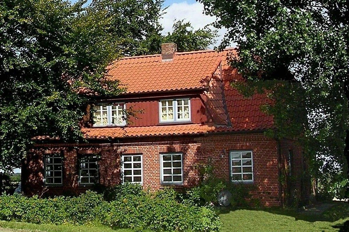 Rotes Ziegelhaus mit Dach und Fenstern, umgeben von Bäumen und Grün.