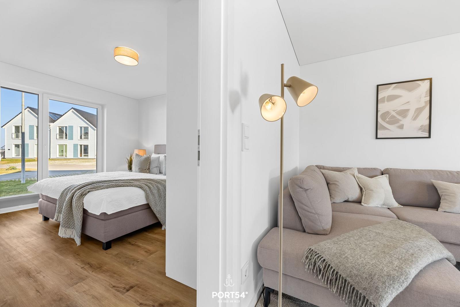 Wohnzimmer mit Sofa und Stehlampe. Nebenraum mit Bett und Fensterblick.