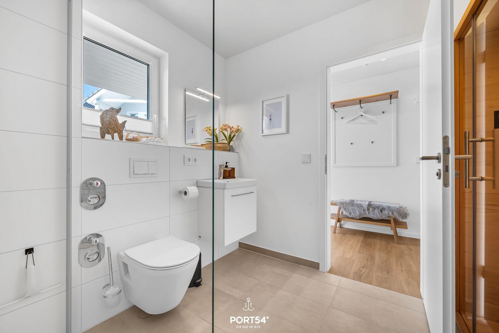 Badezimmer mit Toilette, Waschbecken und Dusche hinter Glas. Nebenraum mit Kleiderständer und Bank.