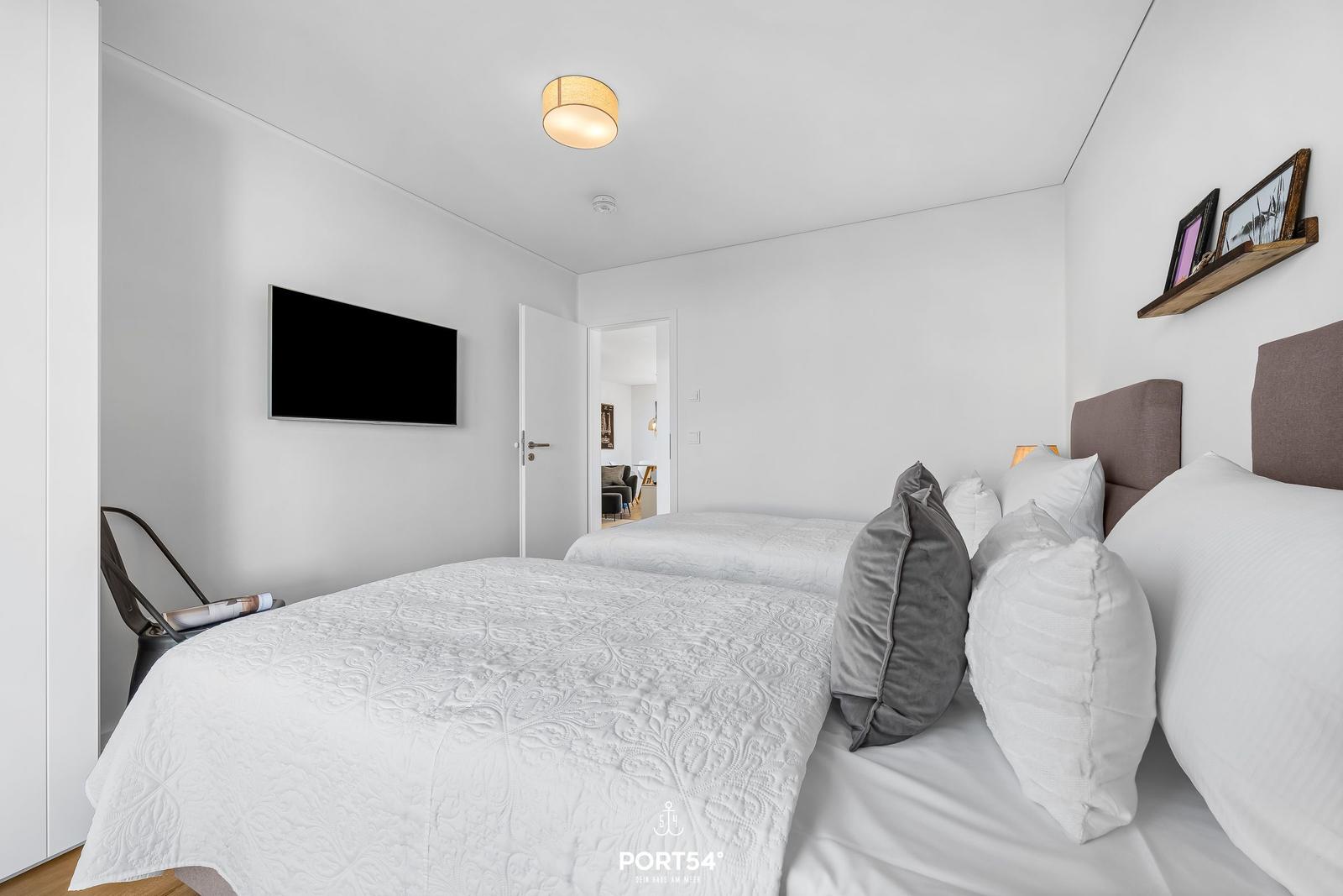 Zwei Betten mit weißen Bettwäsche und Kissen. Wandfernseher und Holzregal an der Wand.