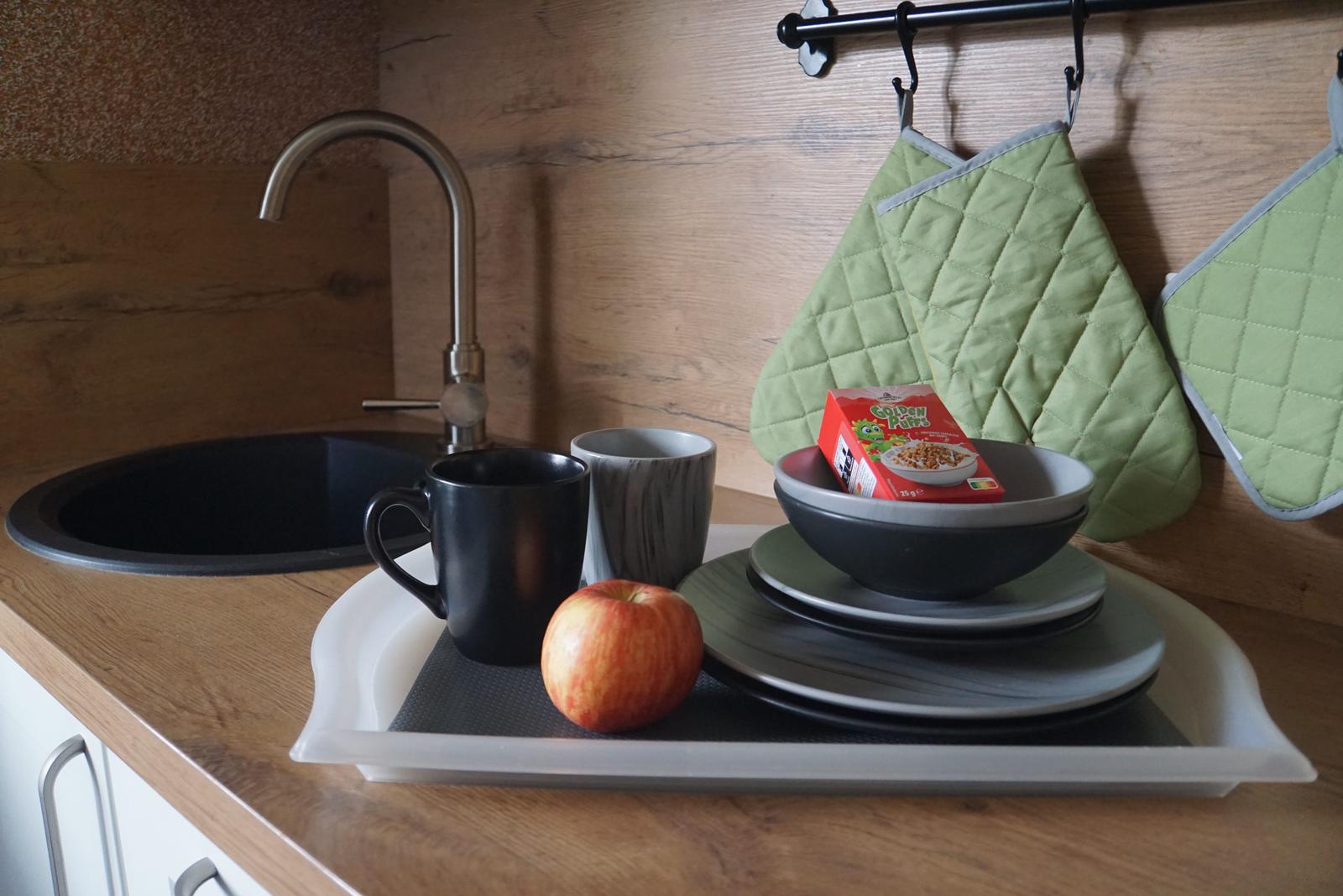 Küchenbereich mit Spüle, Tassen, Teller und Apfel auf Arbeitsplatte.