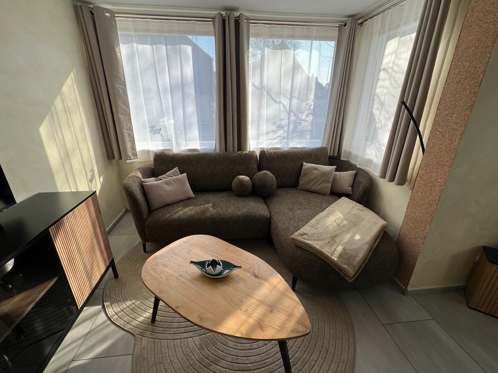 Ein Wohnzimmer mit Ecksofa, Holztisch und Fenstern mit Vorhängen.