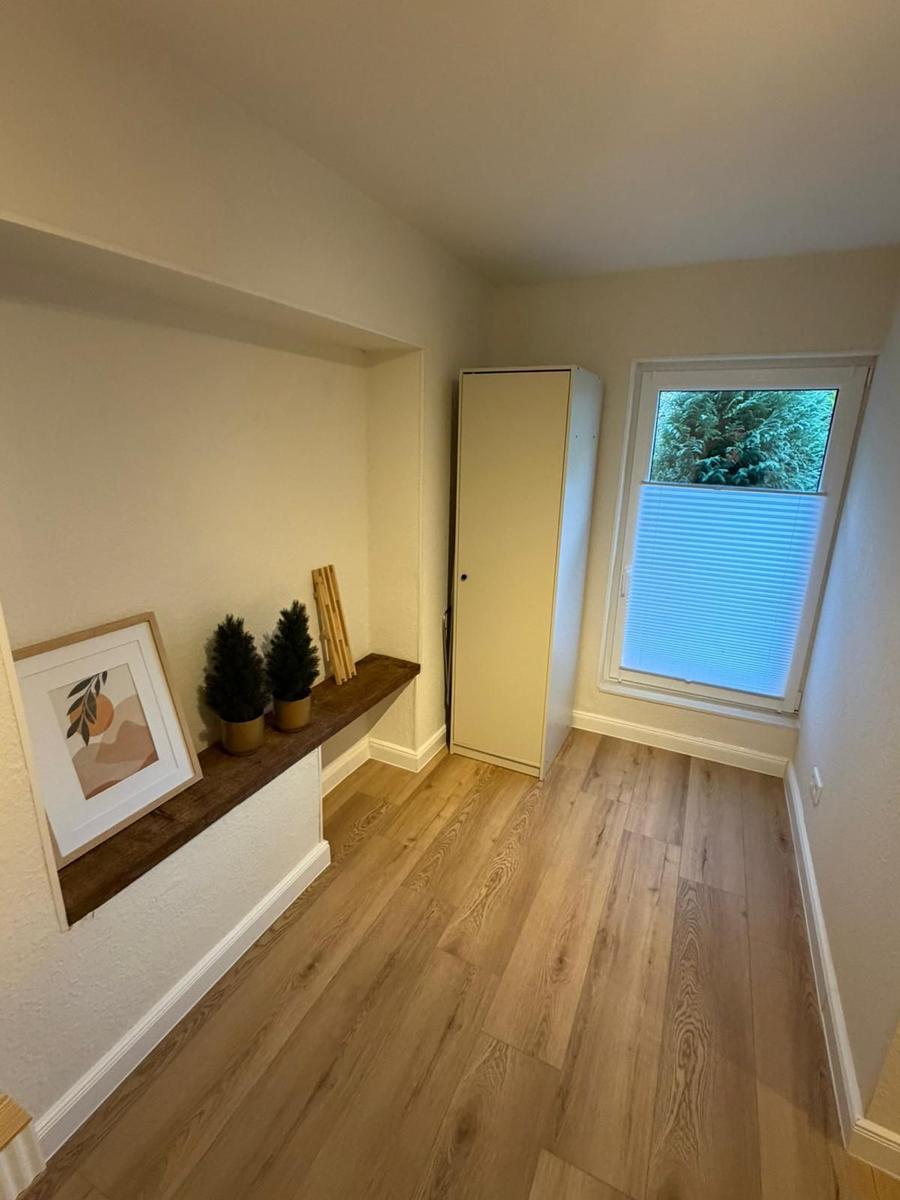 Raum mit Holzboden, Fenster mit Jalousie und kleiner Ablage mit Pflanzen und Bild.