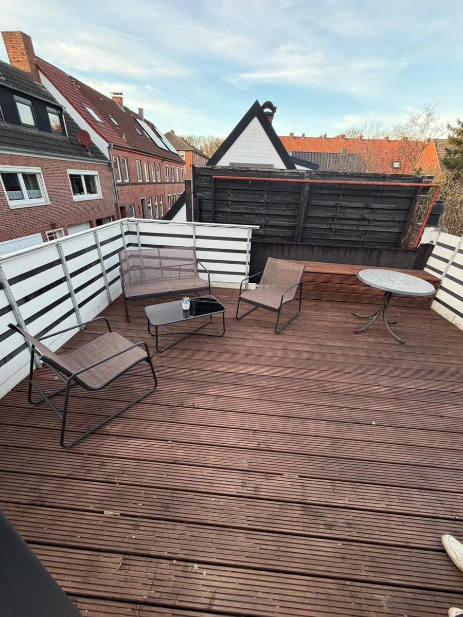 Terrasse mit Holzboden, Sitzmöbeln und Tisch. Umzäunung und Nachbarhäuser im Hintergrund.