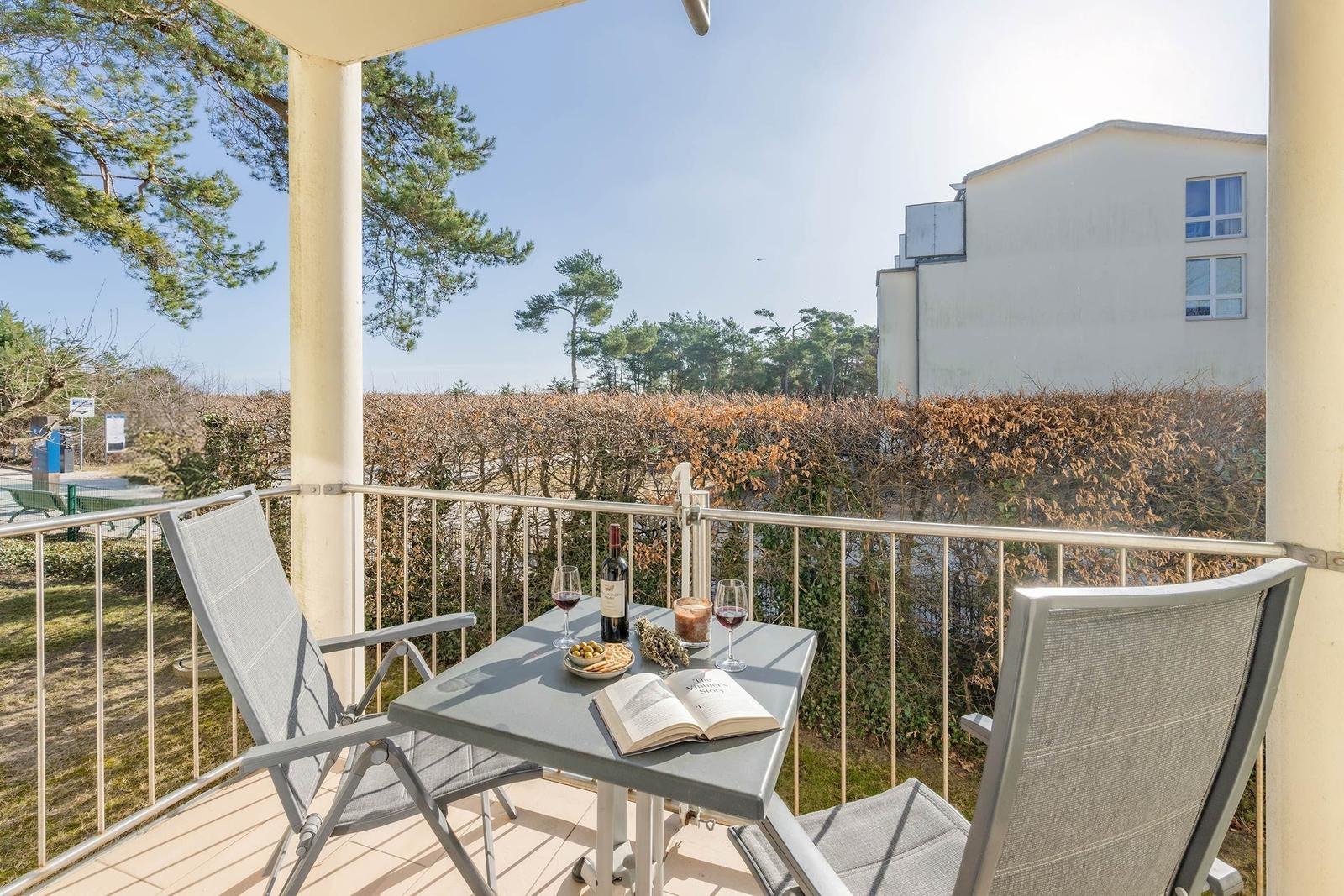 Terrasse mit Tisch, zwei Stühlen, Wein und Buch. Blick auf Bäume und Gebäude.