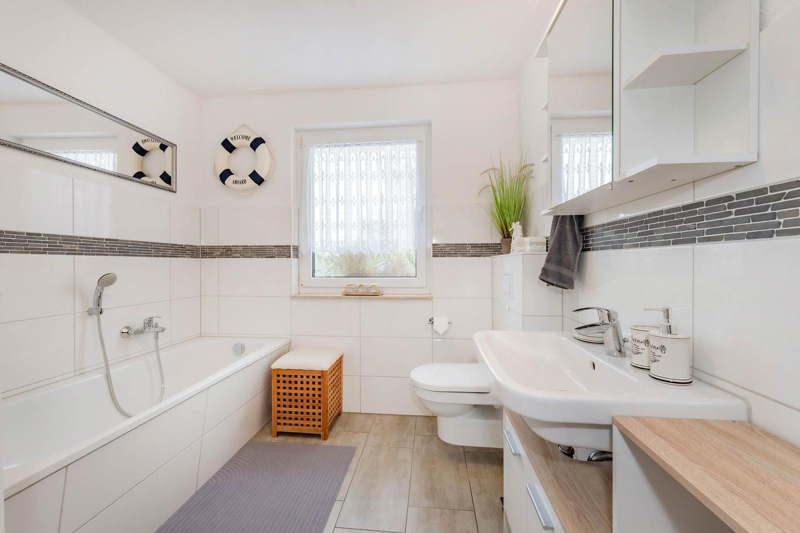 Badezimmer mit Badewanne, Waschbecken und Toilette. Dekorative Elemente wie Rettungsring und Pflanze.