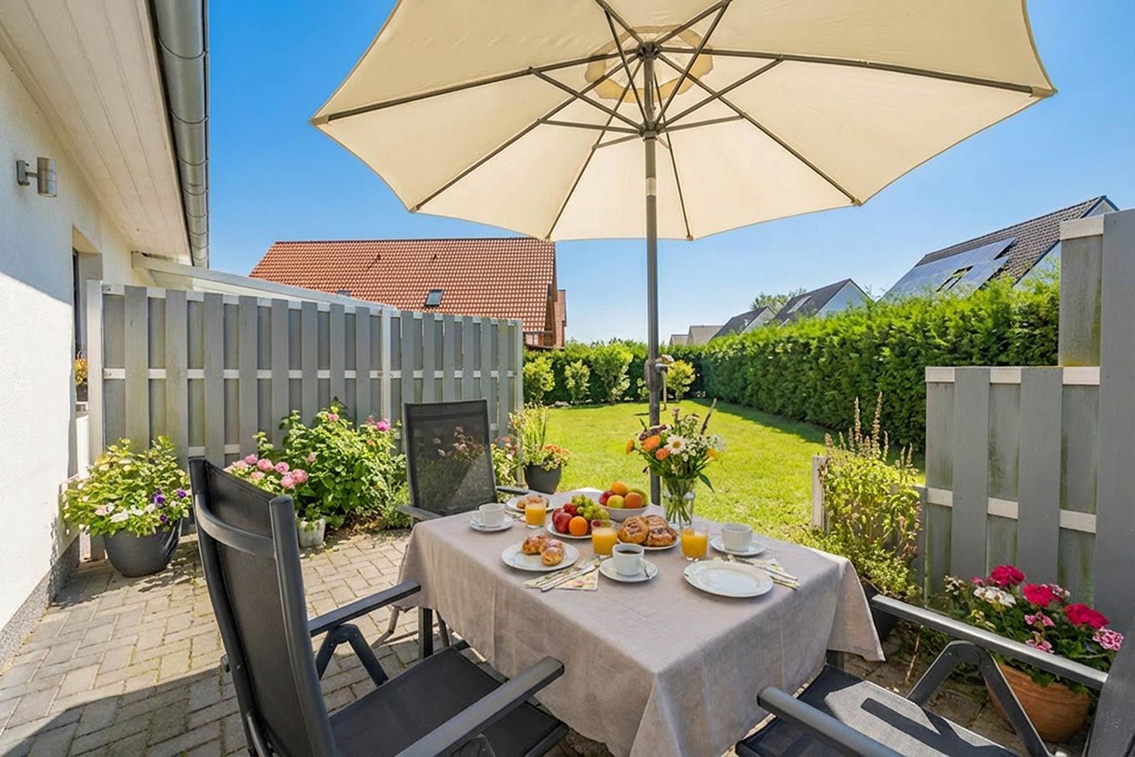 Terrasse mit Tisch, Stühlen und Sonnenschirm. Garten mit Blumen und Rasen.