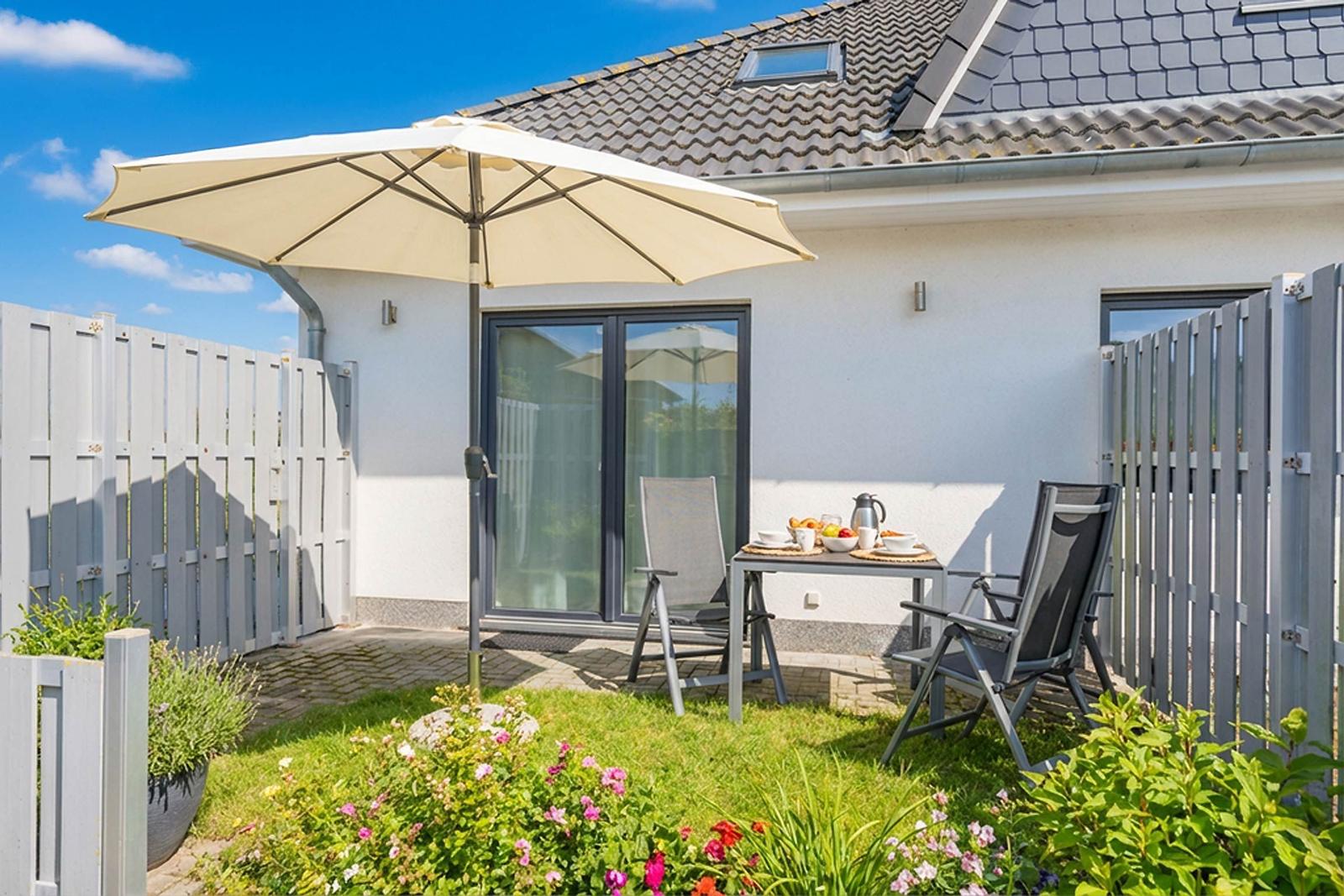 Terrasse mit Tisch, Stühlen und Sonnenschirm. Hintergrund: Haus mit Glastür und Zaun.