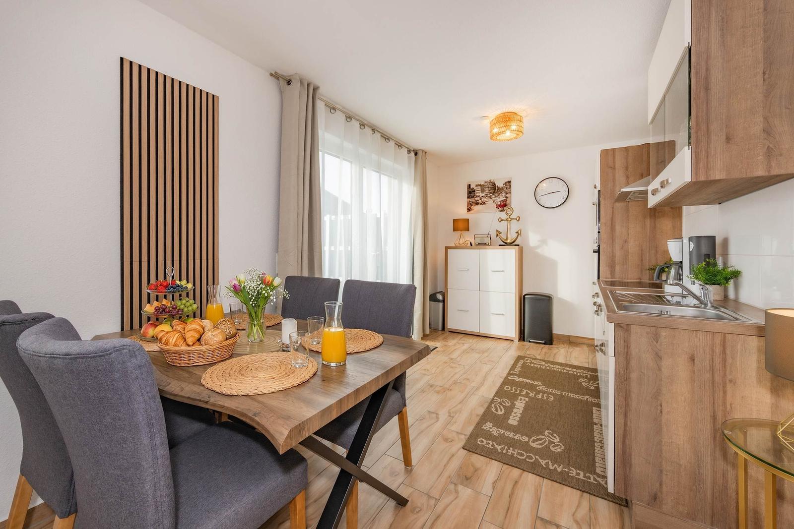 Esszimmer mit Tisch, Stühlen und Küchenbereich. Holzboden und Fenster mit Vorhängen.