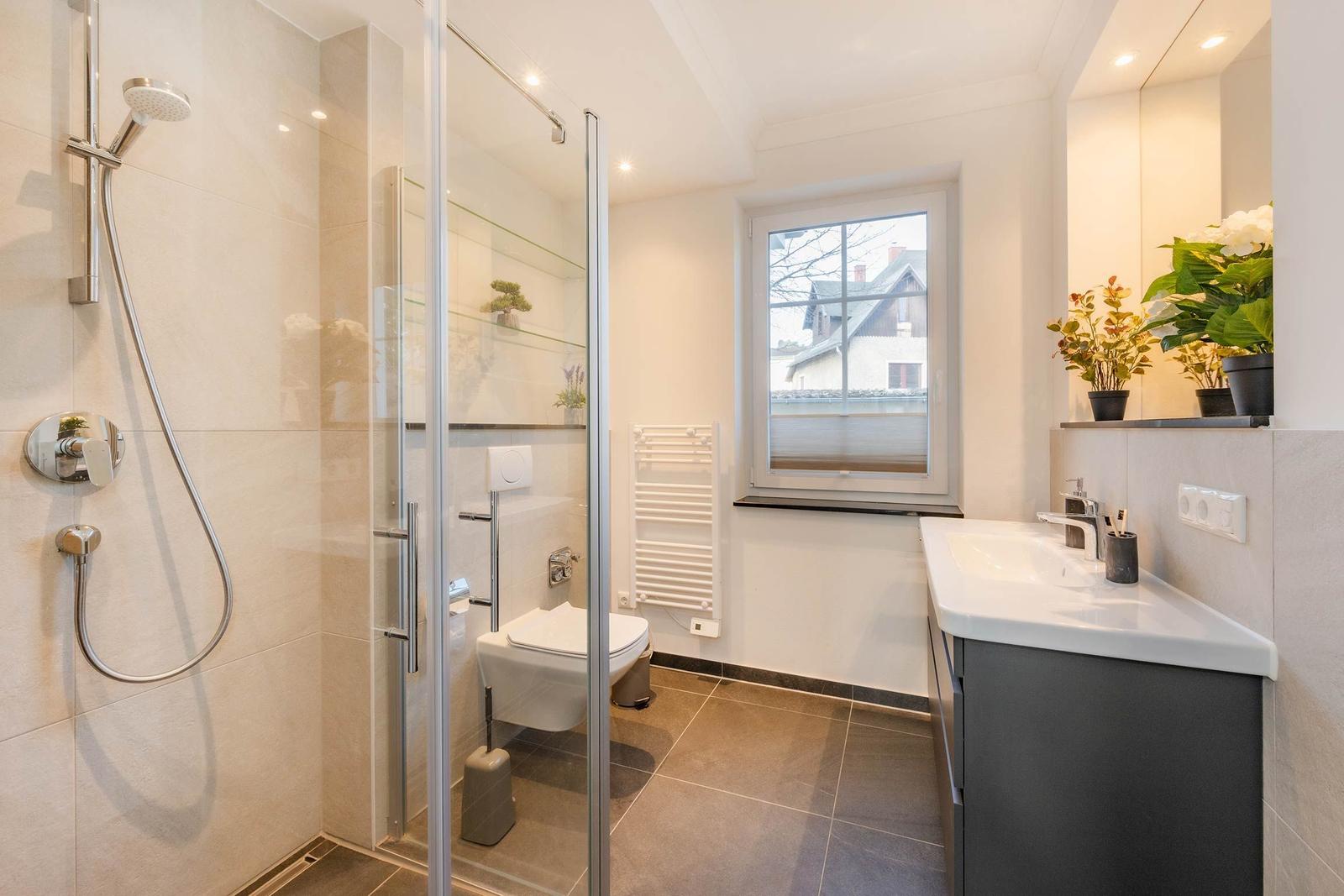 Badezimmer mit Dusche, Toilette und Waschbecken. Fenster mit Blick auf Haus.