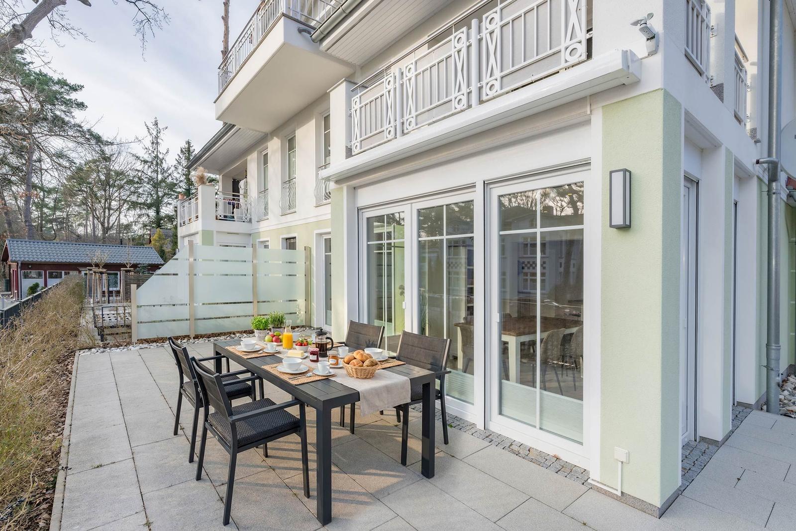 Terrasse mit Tisch und Stühlen, Glasfront und Balkon. Hinterhof mit Holzzaun.