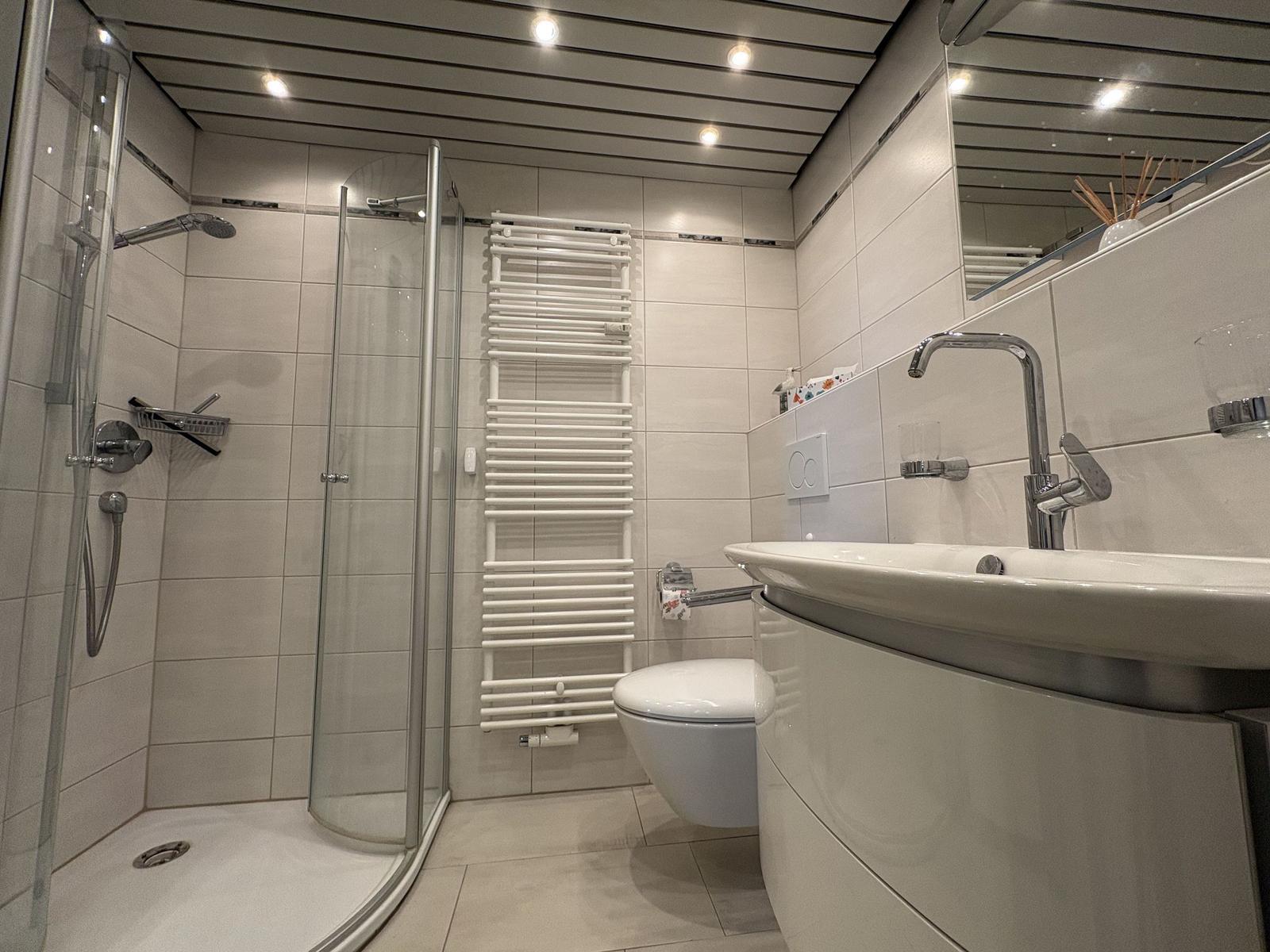 Bad mit Dusche, Waschbecken und Toilette. Weiße Fliesen und moderne Ausstattung.