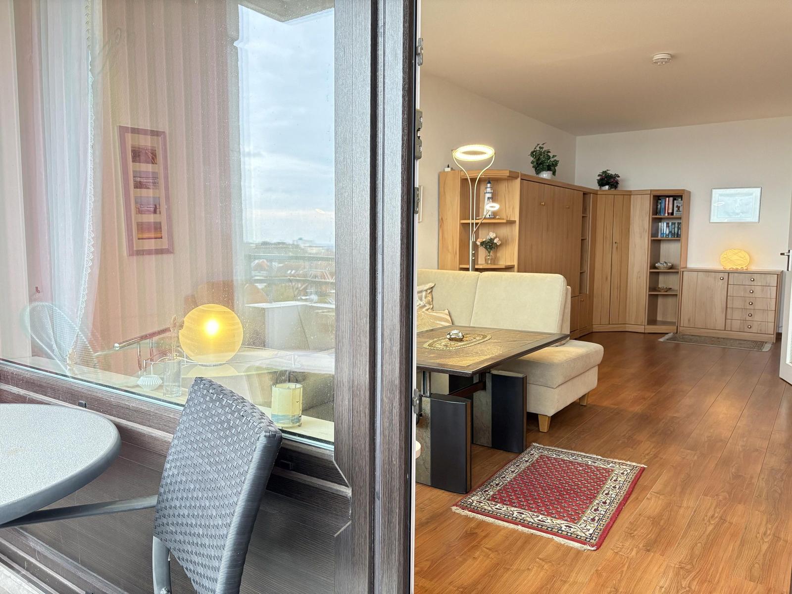 Wohnraum mit Holzboden, Sofa, Tisch und Bücherregal. Durchgang zu Balkon mit Sitzgelegenheit.