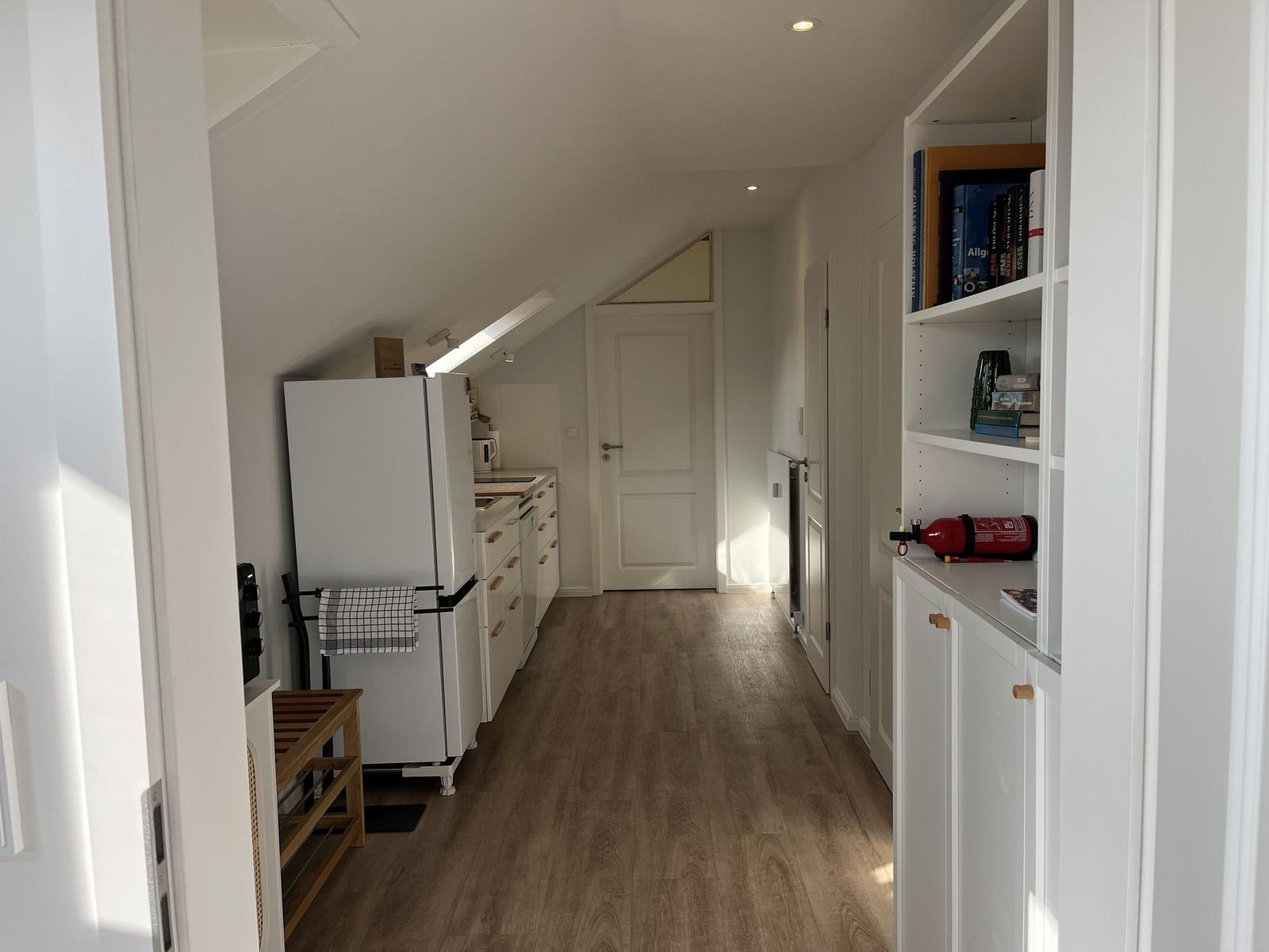 Küche mit Kühlschrank und Holzboden. Regal mit Büchern und Feuerlöscher.