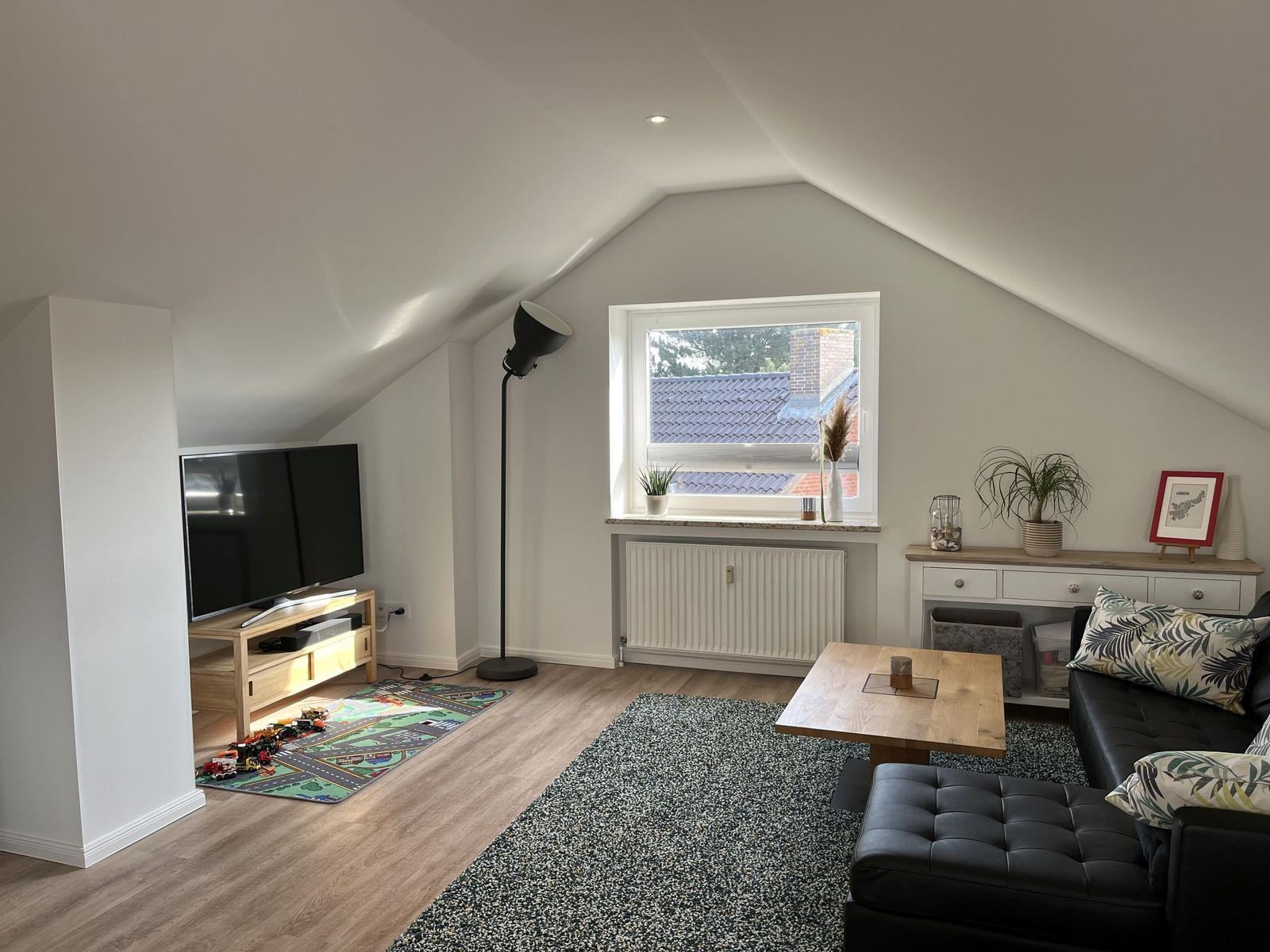 Wohnzimmer mit schwarzem Sofa, Holztisch und Fernseher unter schrägen Dachdecken.