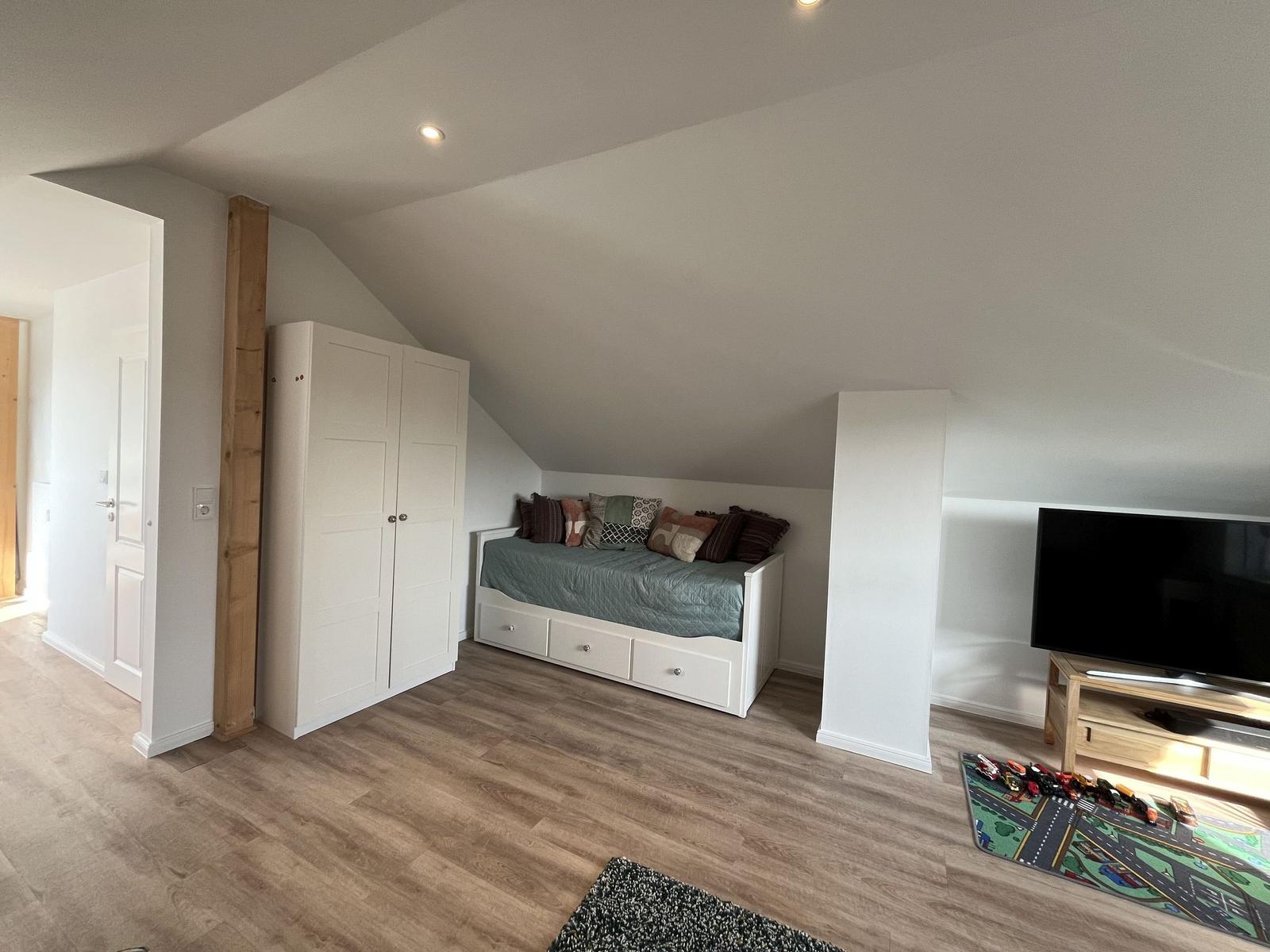 Zimmer mit Dachschrägen, Bett, Kleiderschrank und Fernseher auf Holzstand.
