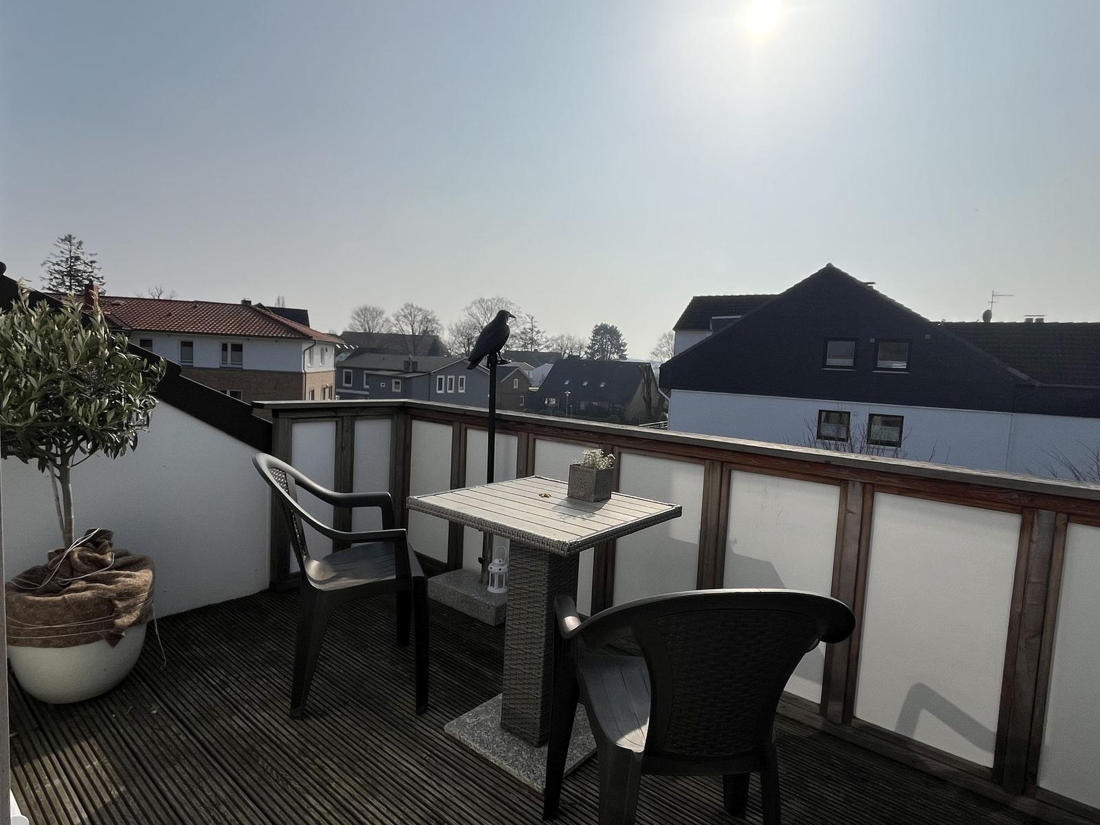 Terrasse mit Tisch, zwei Stühlen und Pflanze. Vogel sitzt auf Pfosten. Blick auf Wohnhäuser.