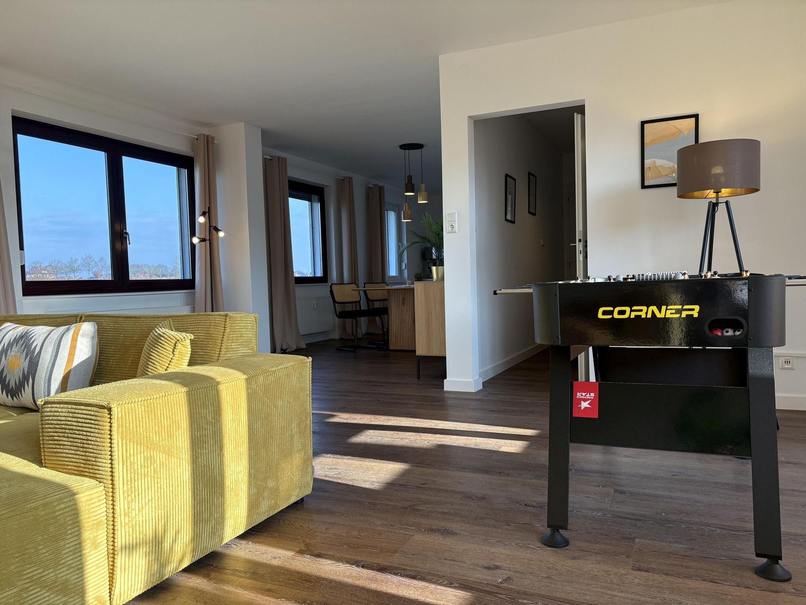 Wohnzimmer mit gelbem Sofa und Tischfußball. Holzboden und große Fenster.