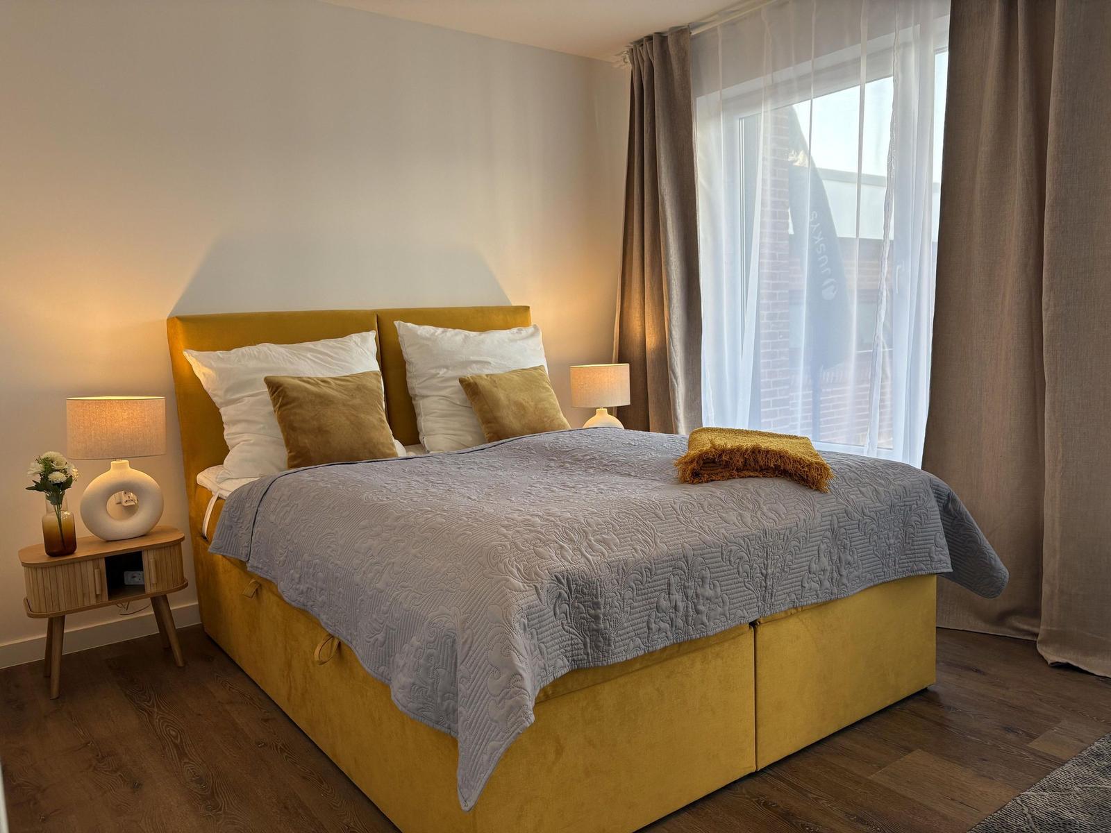 Schlafzimmer mit gelbem Bett, grauer Decke und Holzboden. An den Wänden hängen Vorhänge.