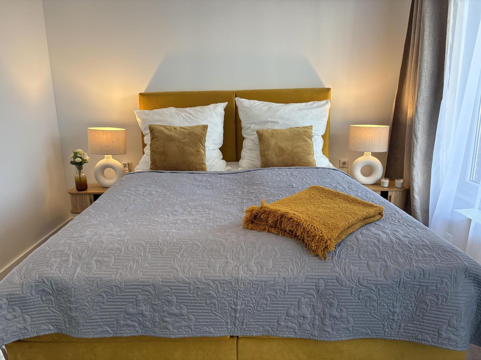 Ein Bett mit gelbem Kopfteil, grauer Decke und Kissen. Beide Nachttische haben Lampen.