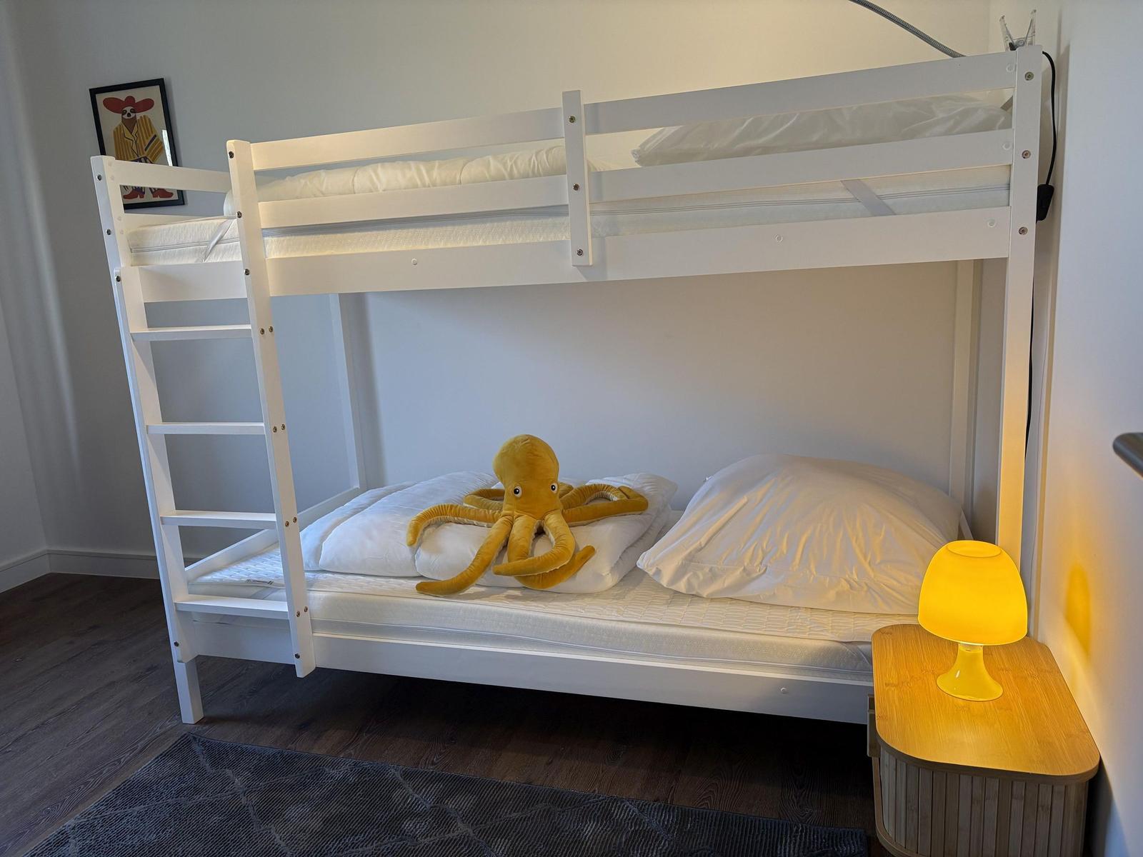 Schlafzimmer mit weißem Doppelstockbett, gelber Tischlampe und Plüschoktopus.
