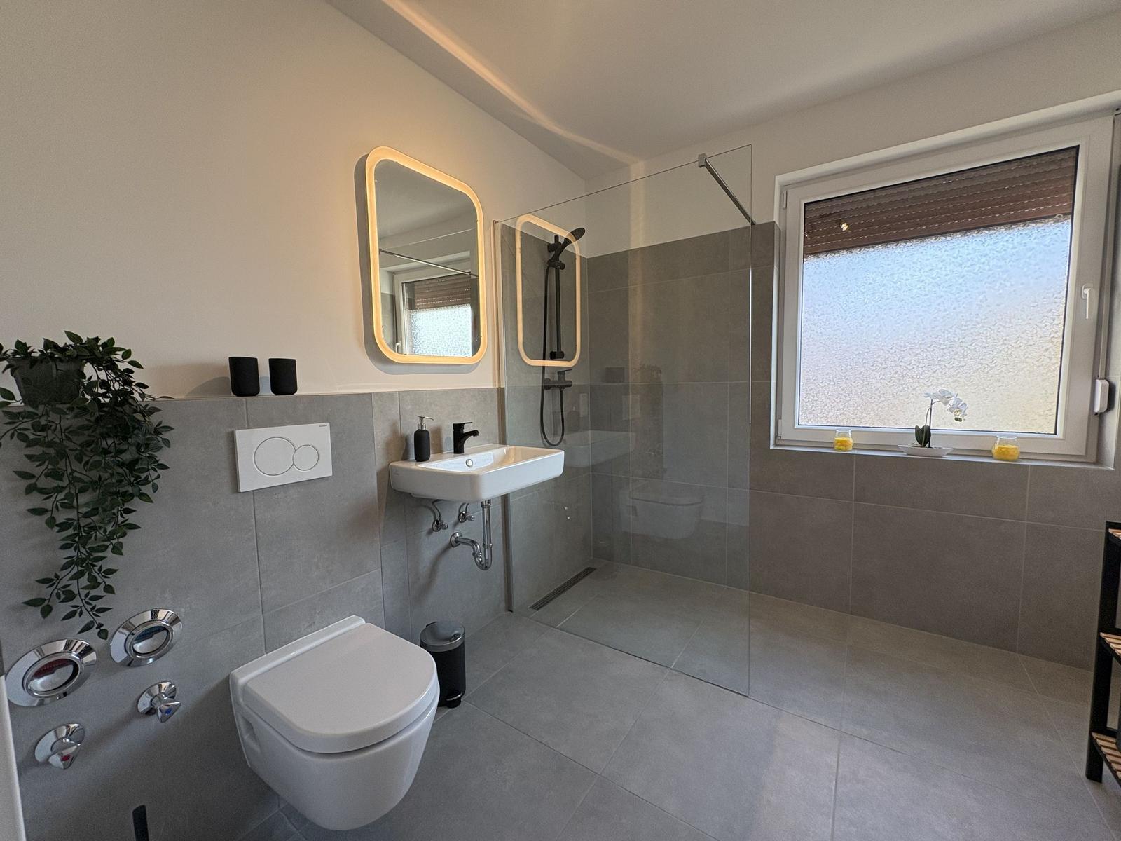 Badezimmer mit Dusche, Waschbecken und Toilette. Fenster mit Rollladen und Pflanzen.