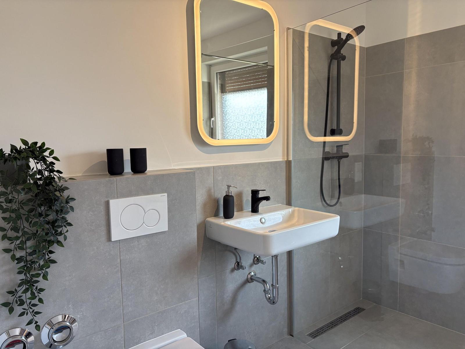 Badezimmer mit Dusche, Waschbecken und Toilette. Graue Fliesen und beleuchtete Spiegel.