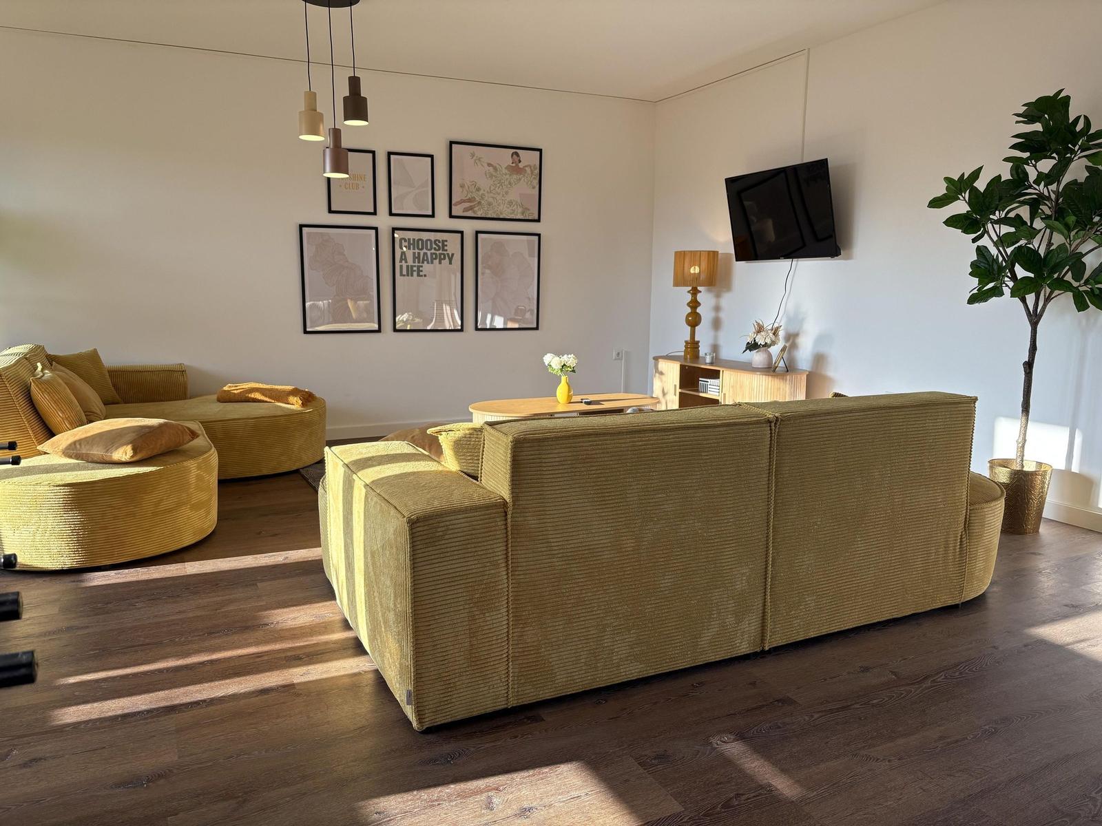 Wohnzimmer mit gelben Sitzmöbeln, Wandbildern und Fernseher.