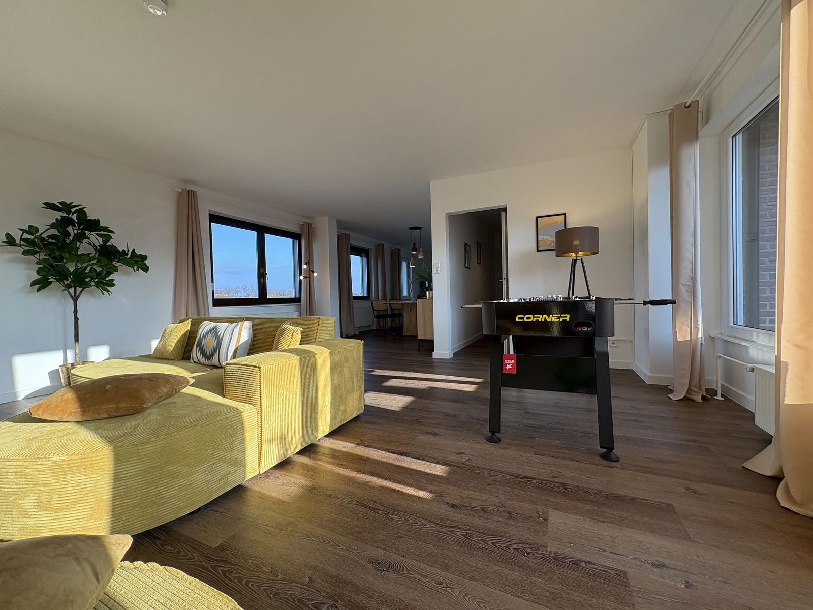 Wohnbereich mit gelbem Sofa und Tischfußball. Holzboden und große Fenster.