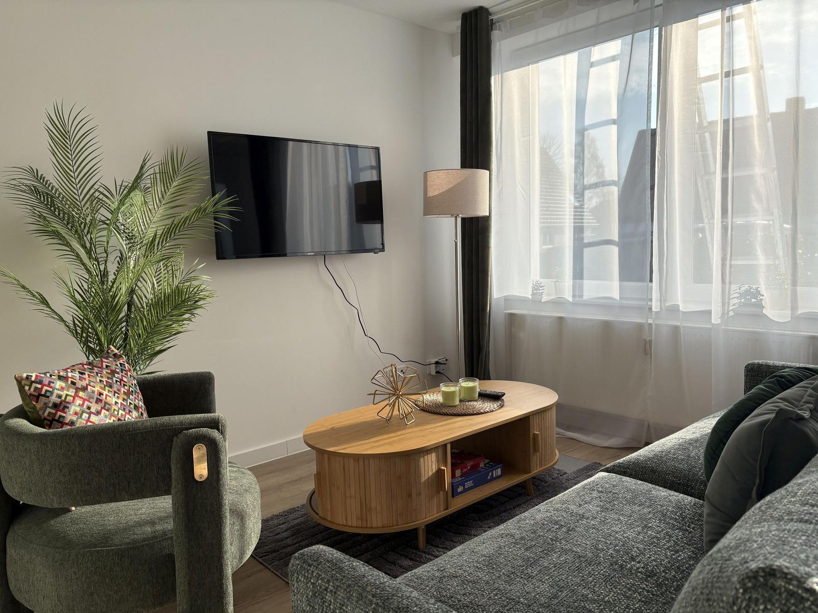 Wohnzimmer mit Sofa, Sessel, Fernseher und Pflanze.