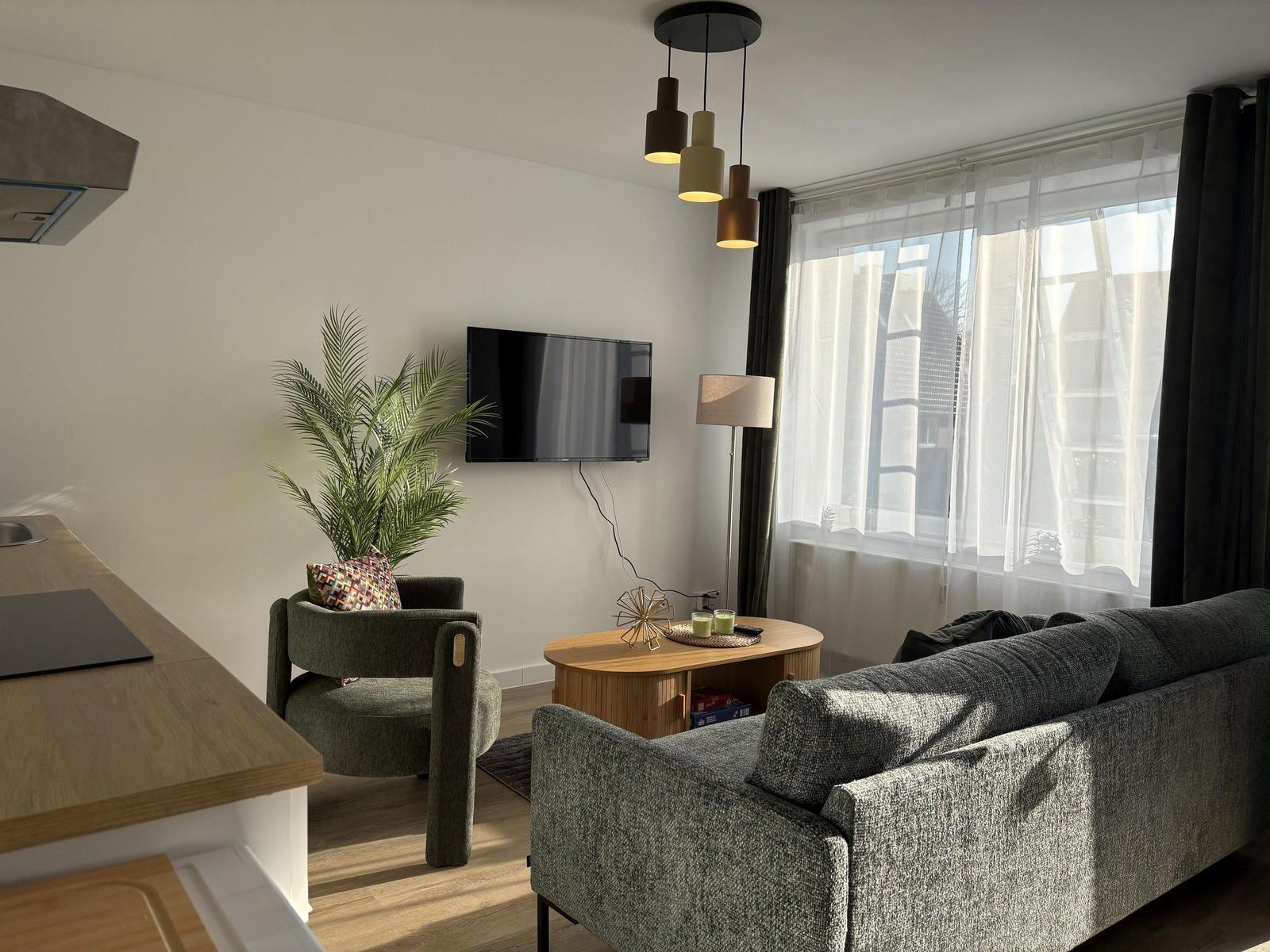 Wohnbereich mit Sofa, Sessel, TV und Fenster mit Gardinen.