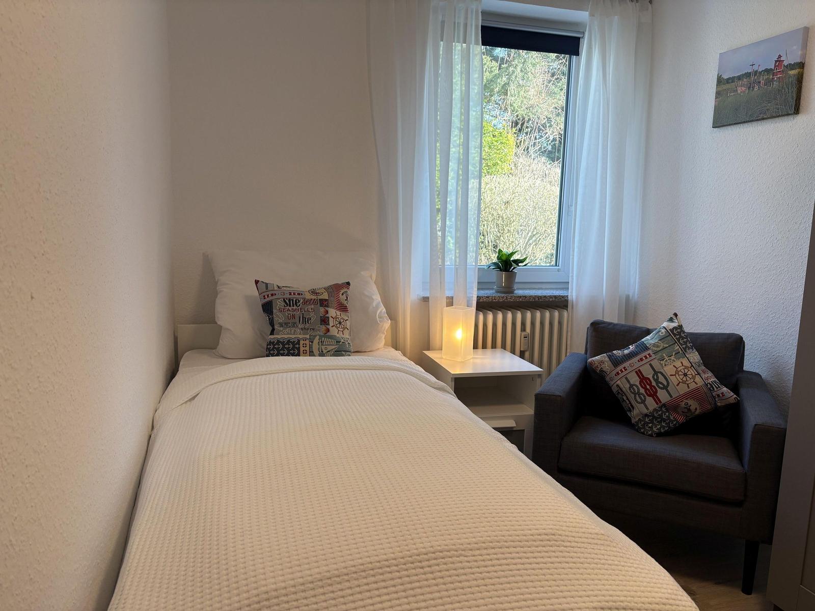 Ein Schlafzimmer mit Bett, Sessel und Fenster. Auf dem Fensterbrett steht eine Pflanze.