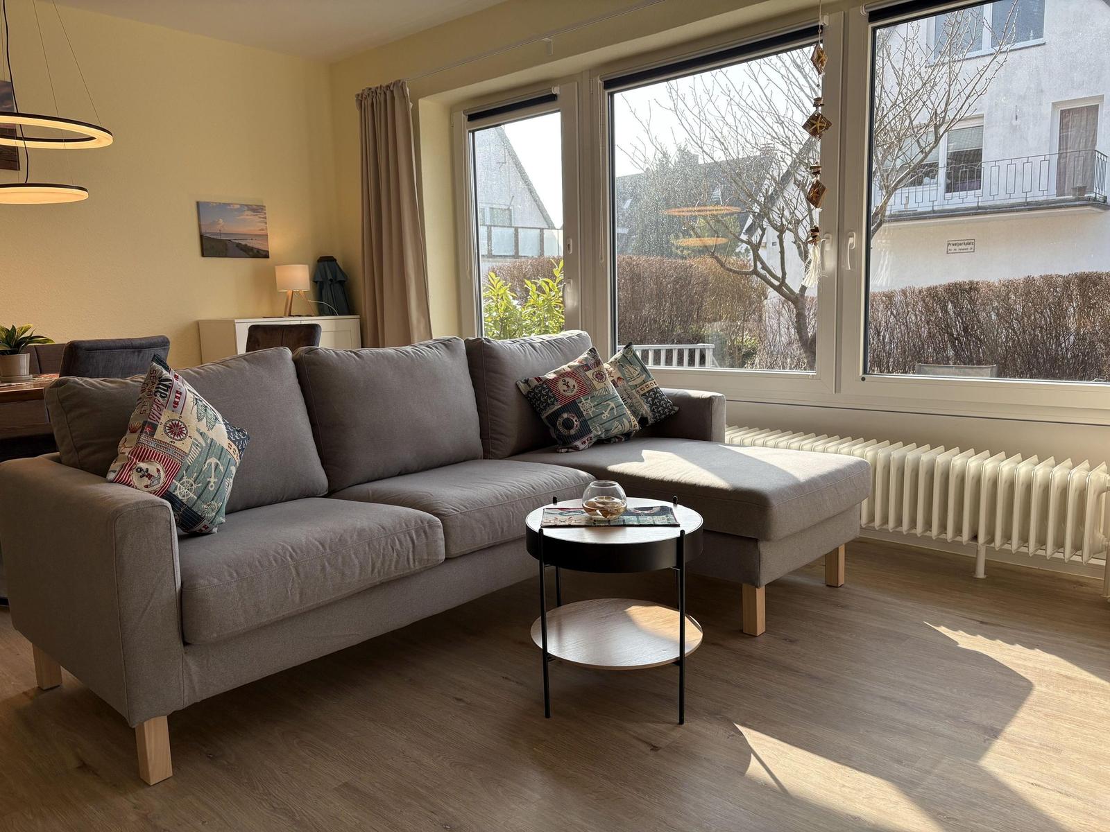 Ein Wohnzimmer mit grauem Sofa, kleinem Tisch und großen Fenstern.