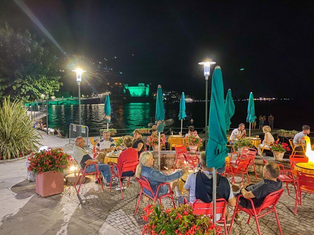 Nachtansicht einer Terrasse mit Gästen, roten Stühlen, grünen Sonnenschirmen und Blick auf einen beleuchteten See.