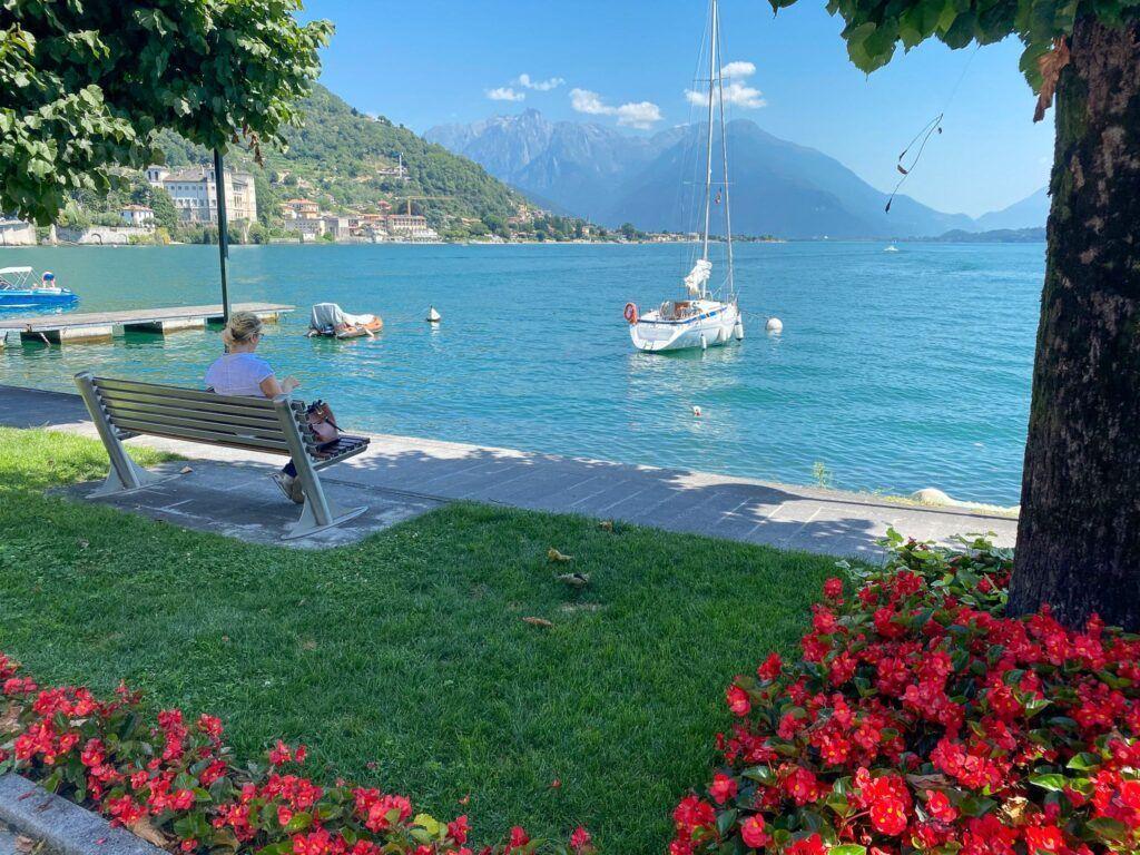 Eine Person sitzt auf einer Bank am Ufer eines Sees mit Booten und Bergen im Hintergrund.