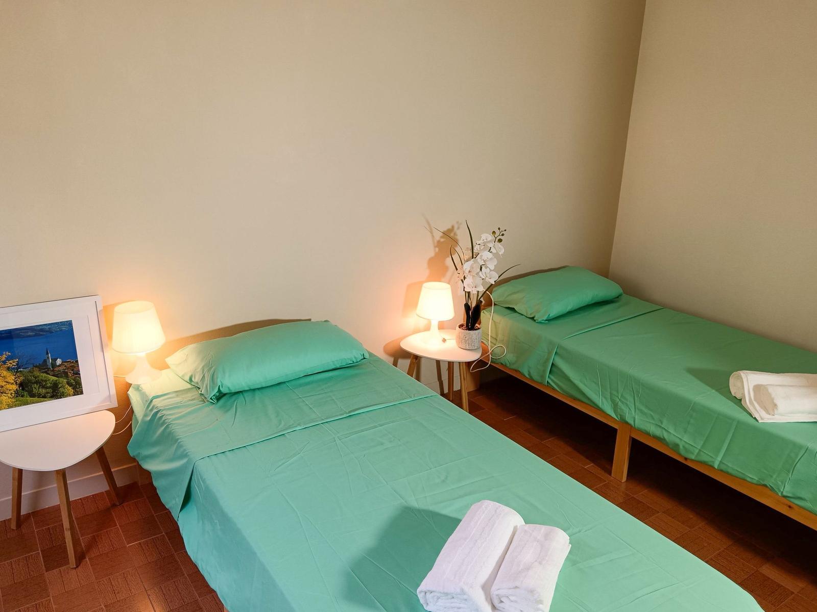 Zwei Betten mit grünen Bettwäschen, Nachttische mit Lampen und Blumen.