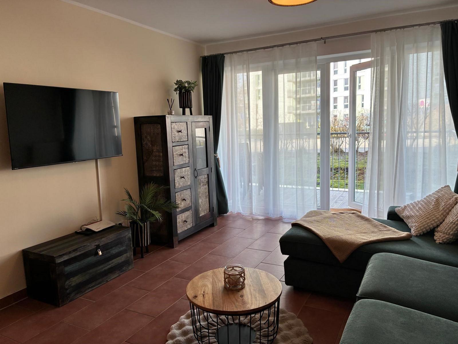 Wohnzimmer mit Sofa, TV und Balkon. Holztisch und Schrank sind sichtbar.