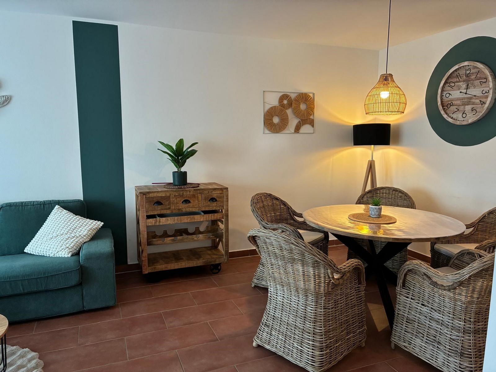 Essbereich mit runden Tisch und Stühlen. Sofa, Wanduhr und Beleuchtung sind sichtbar.