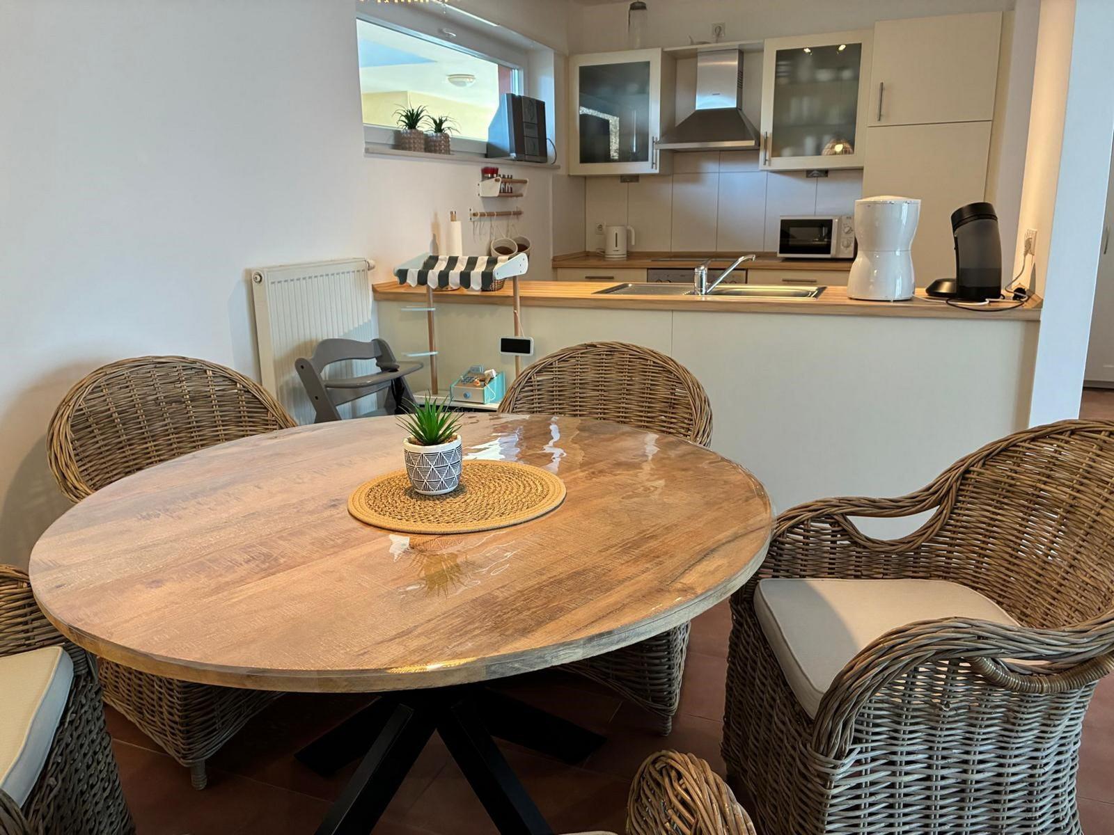 Runder Holztisch mit Strohstühlen und Küchenbereich im Hintergrund.