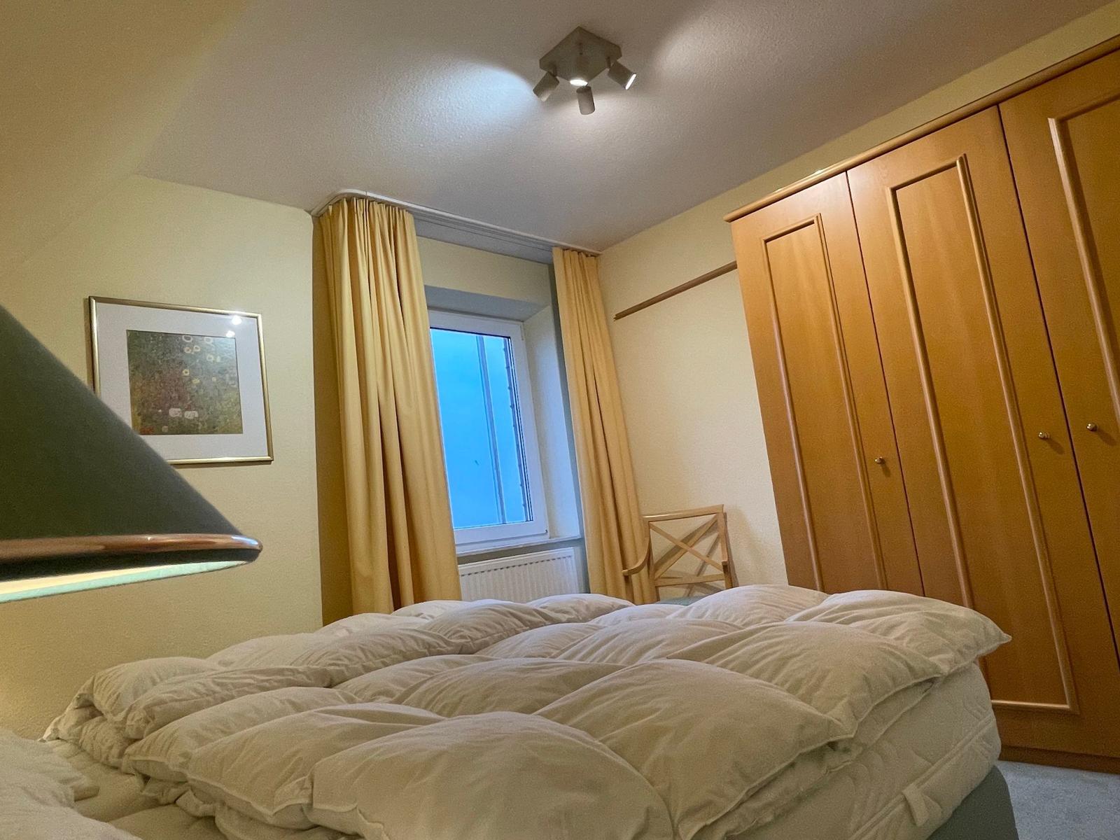 Schlafzimmer mit Bett, Kleiderschrank und Fenster mit Vorhängen.