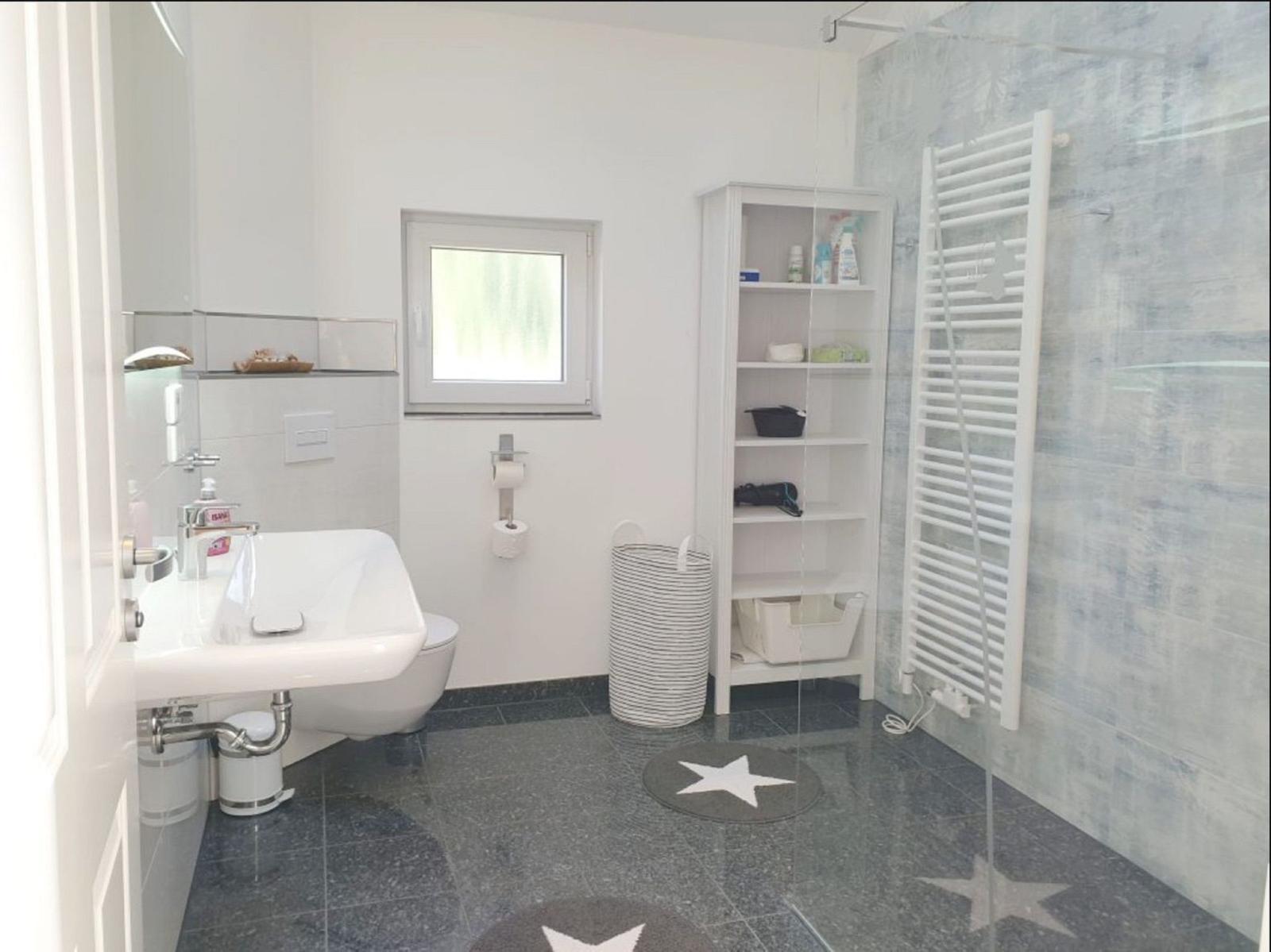 Badezimmer mit Dusche, Waschbecken und Regal. Fliesenboden und Fenster.