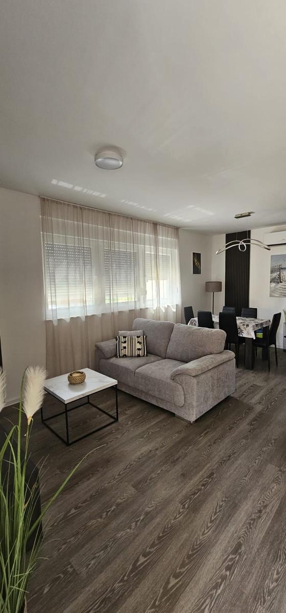 Wohnzimmer mit Sofa, Tisch und Essbereich. Holzboden und Fenster mit Vorhängen.