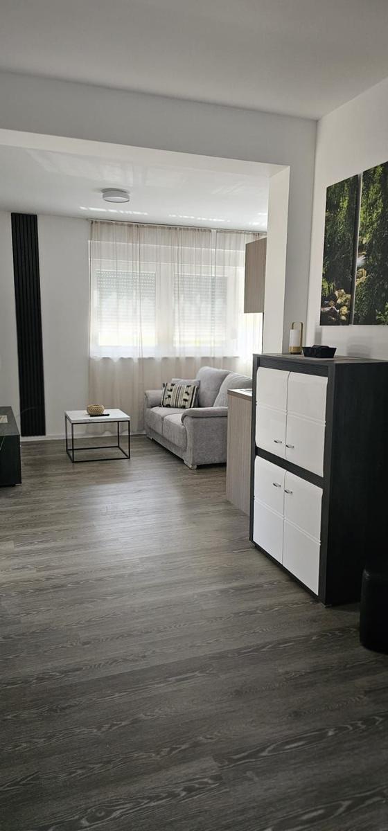 Wohnzimmer mit grauem Sofa, Holzboden und Schrank. Fenster mit weißen Vorhängen.