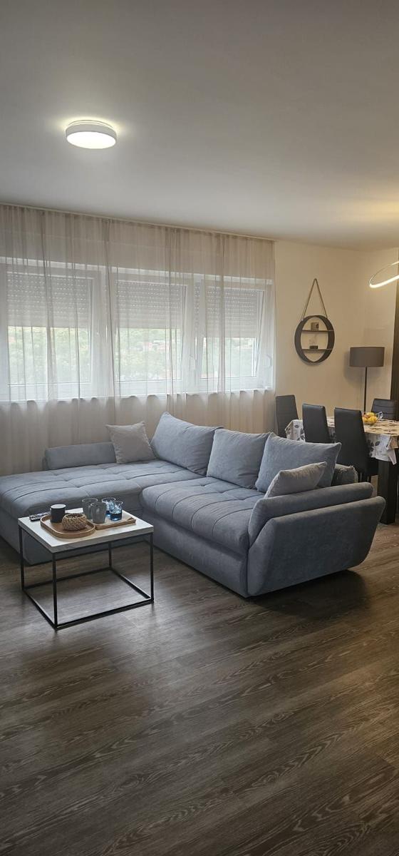 Großer Wohnbereich mit grauem Sofa und Holzboden. Fenster mit weißen Vorhängen.