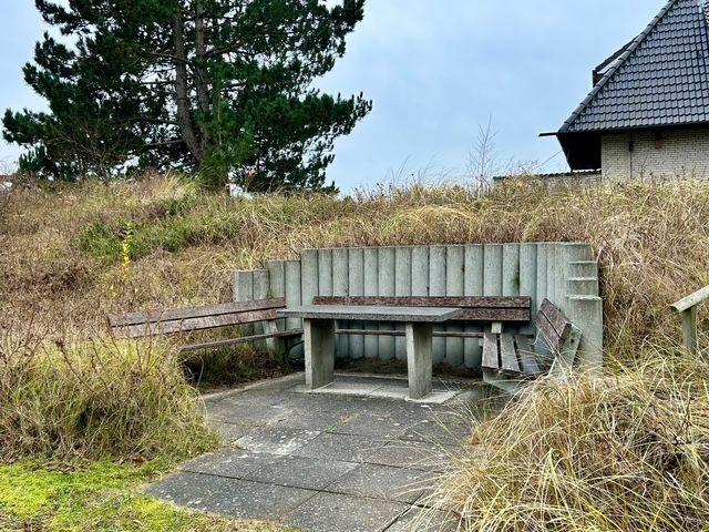 Eine Bank aus Beton und Holz auf einem Steinweg in einer grasbewachsenen Umgebung mit einem Haus im Hintergrund.