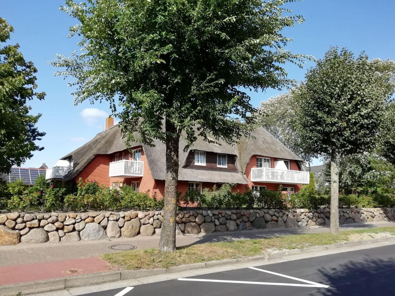 Rotes Haus mit Strohdach und weißen Balkonen. Steinmauer und Bäume umgeben die Fassade.