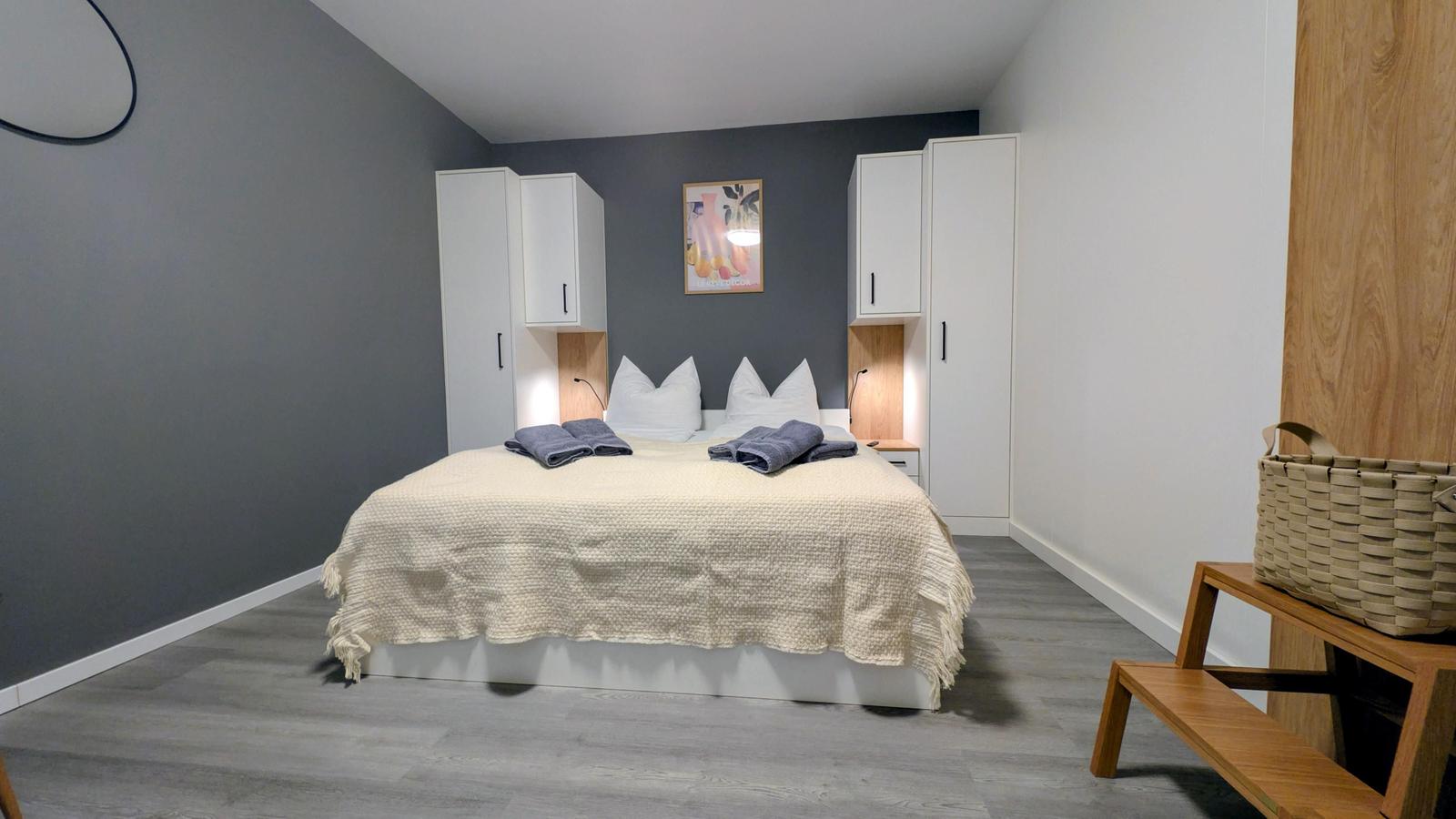Schlafzimmer mit Bett, Schränken und Holzbank. Wandfarbe grau, Boden hellgrau.