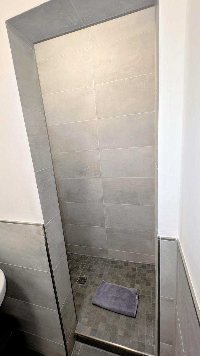 Dusche mit grauen Fliesen und dunklem Boden, ein Handtuch liegt darauf.